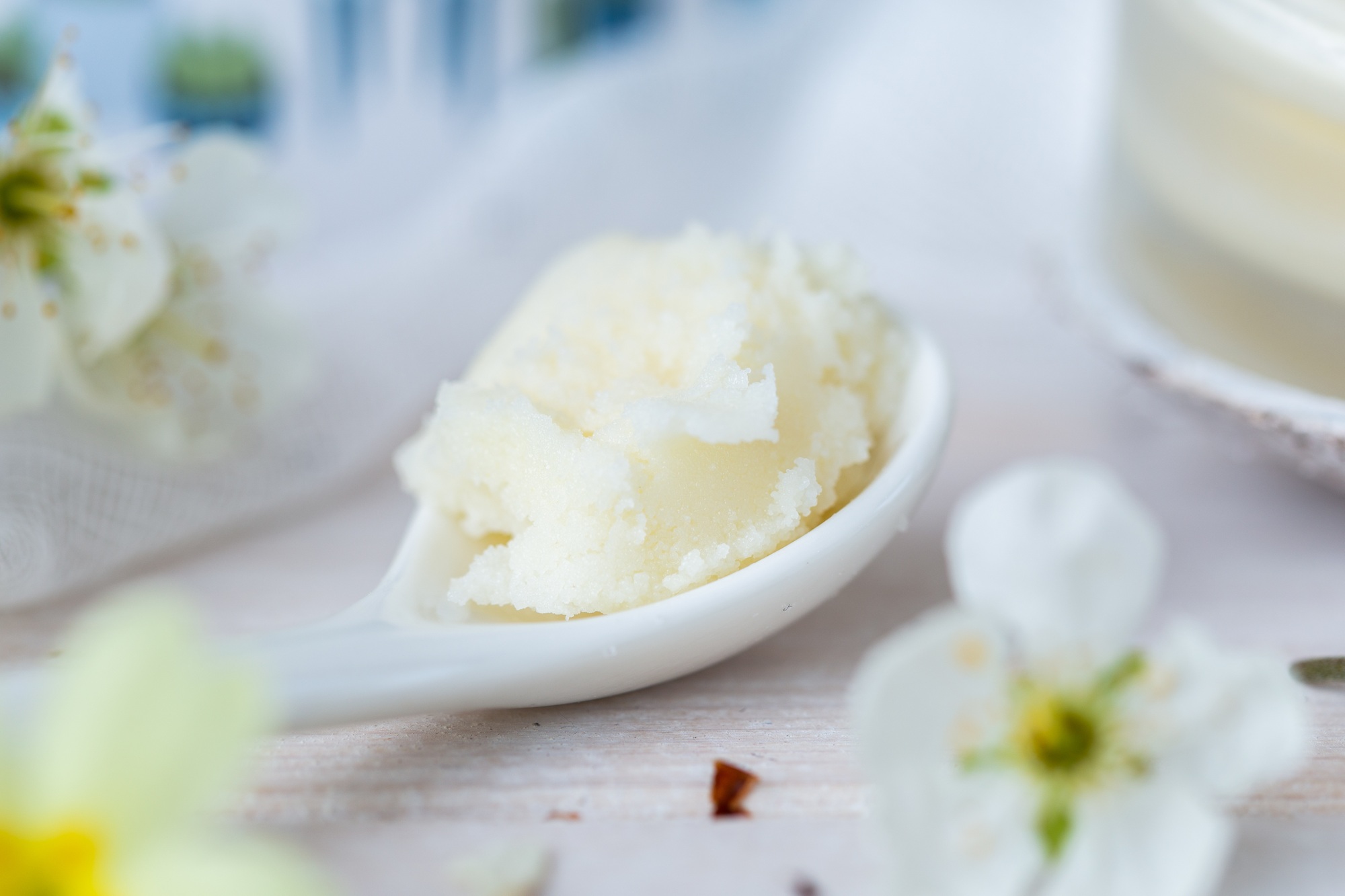 Selective focus shot of a spoonful of shea butter and some white flowers on a white table top
